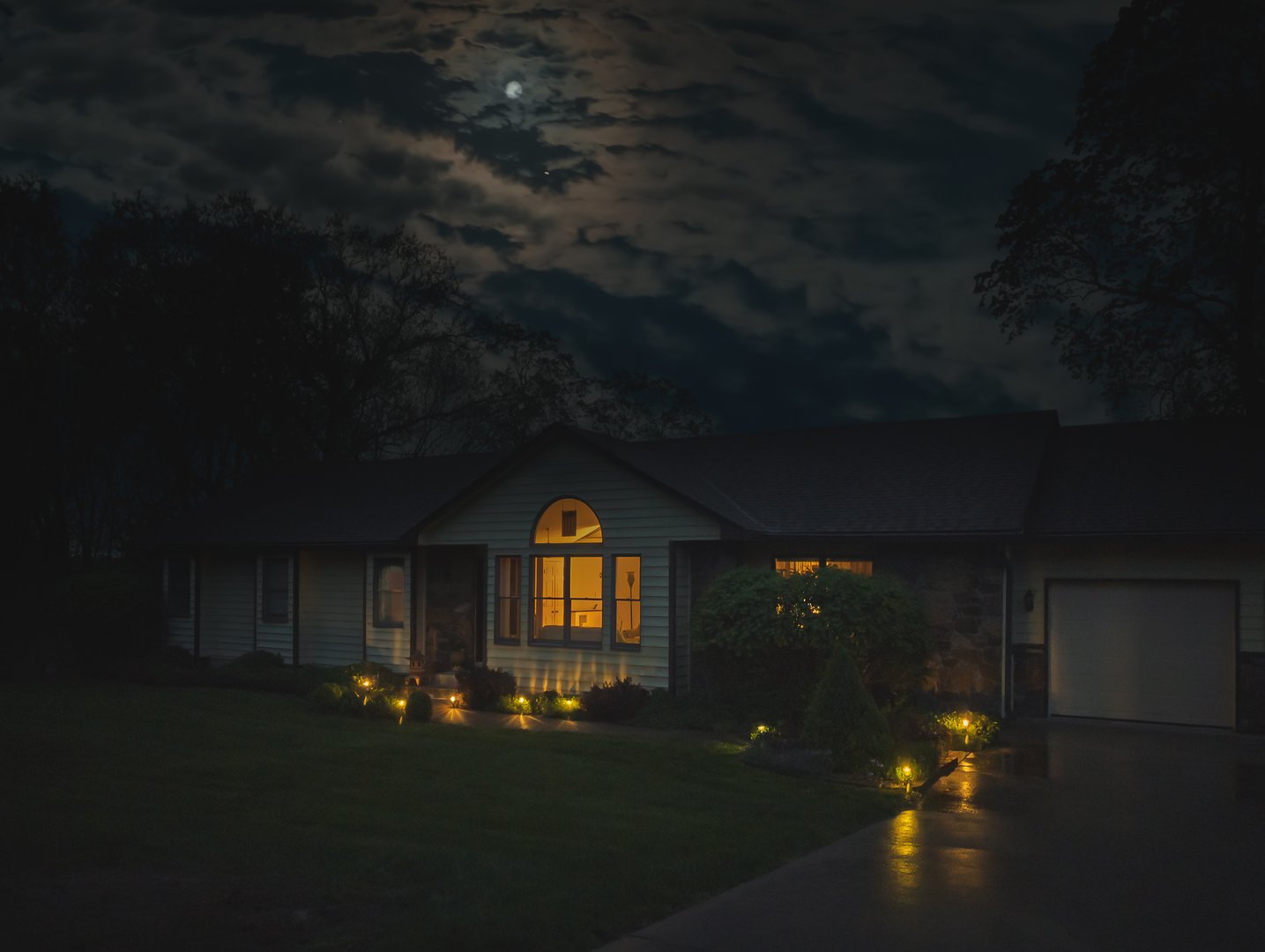 Suburban Midwestern house after rain with moon peeking through clouds; lights in house reflect in wet pavement