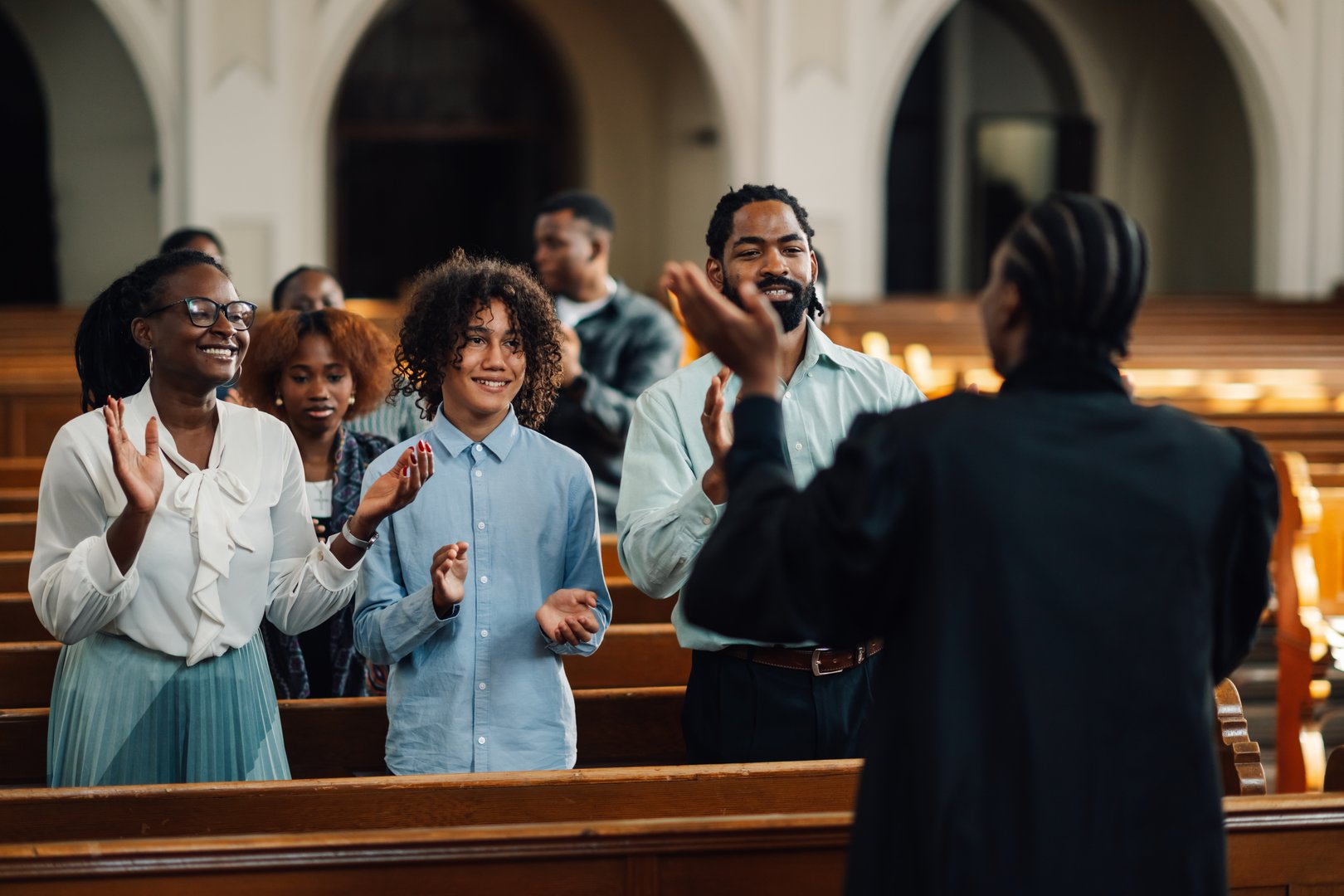 Diverse group of churchgoers are clapping along to their preacher, who is leading the service. The parishioners are smiling and engaged in the service