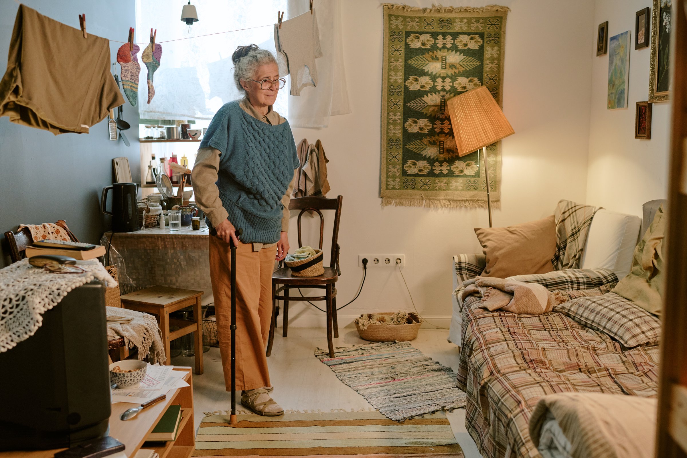 Senior Caucasian woman standing with cane in modest living room, wearing glasses and smiling gently, surrounded by simple furnishings and household items, showing signs of poverty