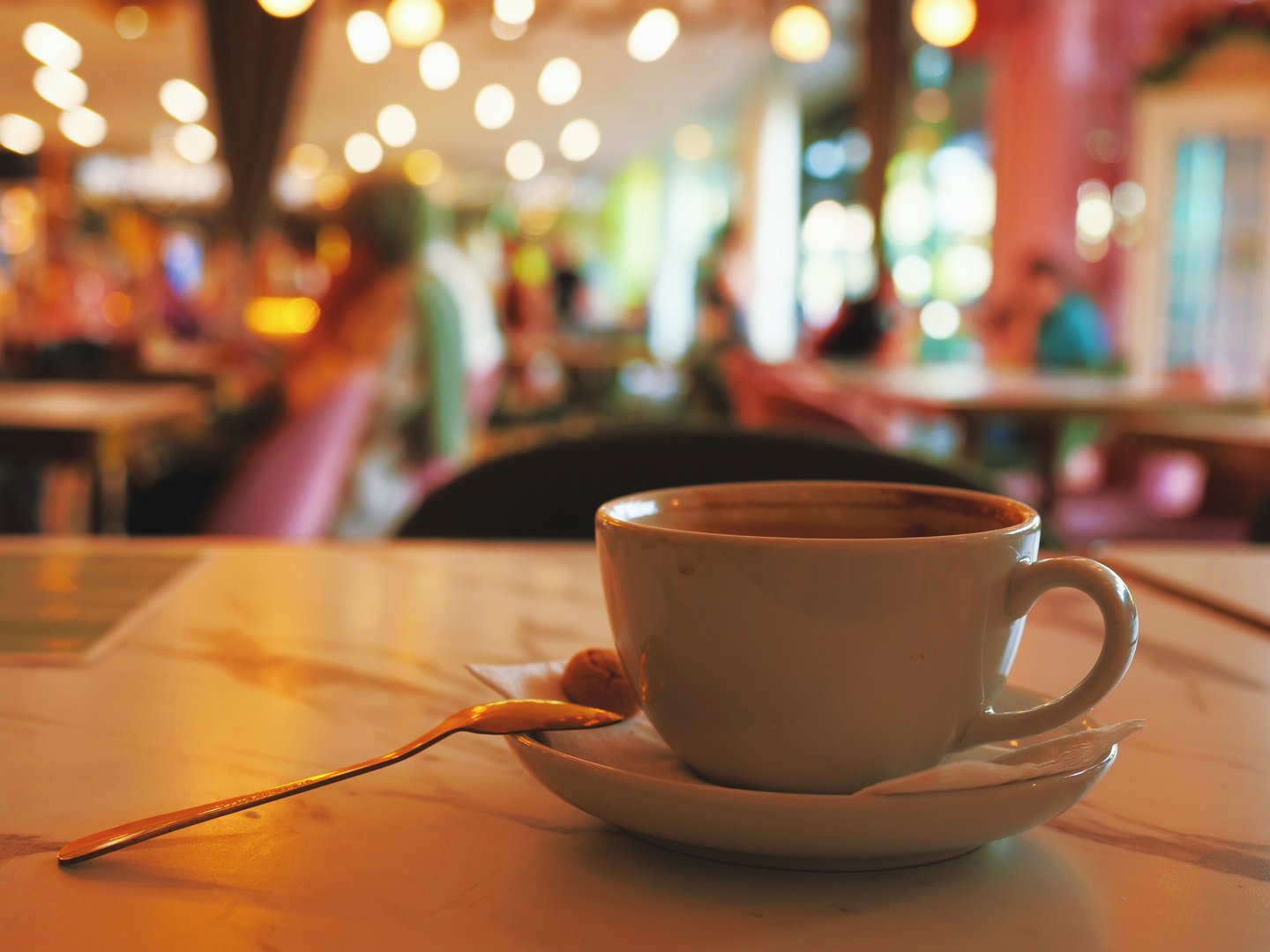 White coffee cup with saucer and spoon in cozy café – dark brew, biscuit, blurred background with people, warm lighting, green and pink decor, relaxing moment, urban lifestyle, public space