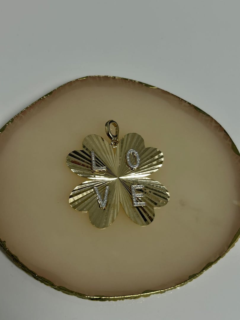 Gold four-leaf clover pendant with the word LOVE engraved, placed on a round, gold-rimmed dish.