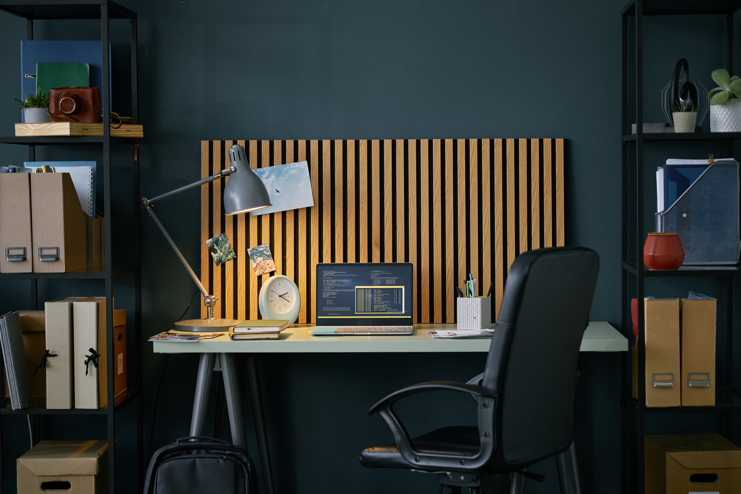 Modern home office setup with a wooden backdrop, a desk lamp, and a laptop on a white table surrounded by black shelves filled with books and storage boxes