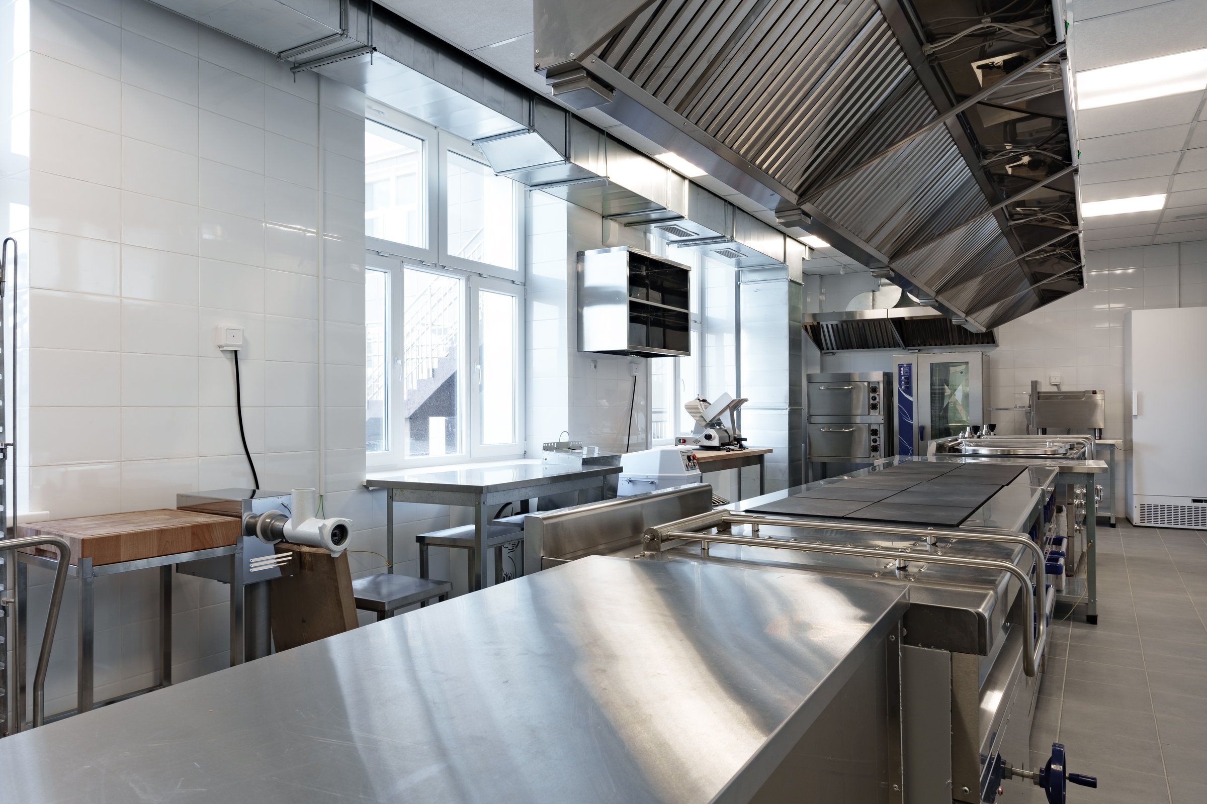 A spacious commercial kitchen features stainless steel surfaces, large ventilation hoods, and ample natural light streaming through multiple windows.
