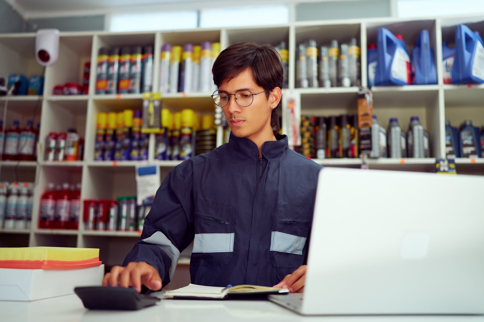 Focused mechanic using calculator and laptop, managing inventory and finances in a busy auto shop, surrounded by shelves of car parts and supplies