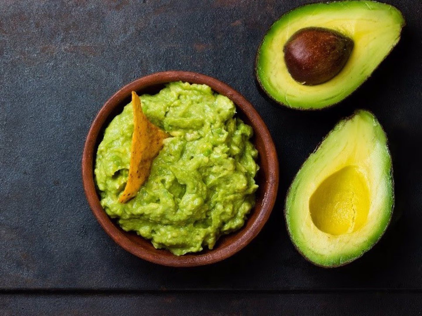 Bowl of guacamole with a tortilla chip next to a halved avocado on a dark surface.