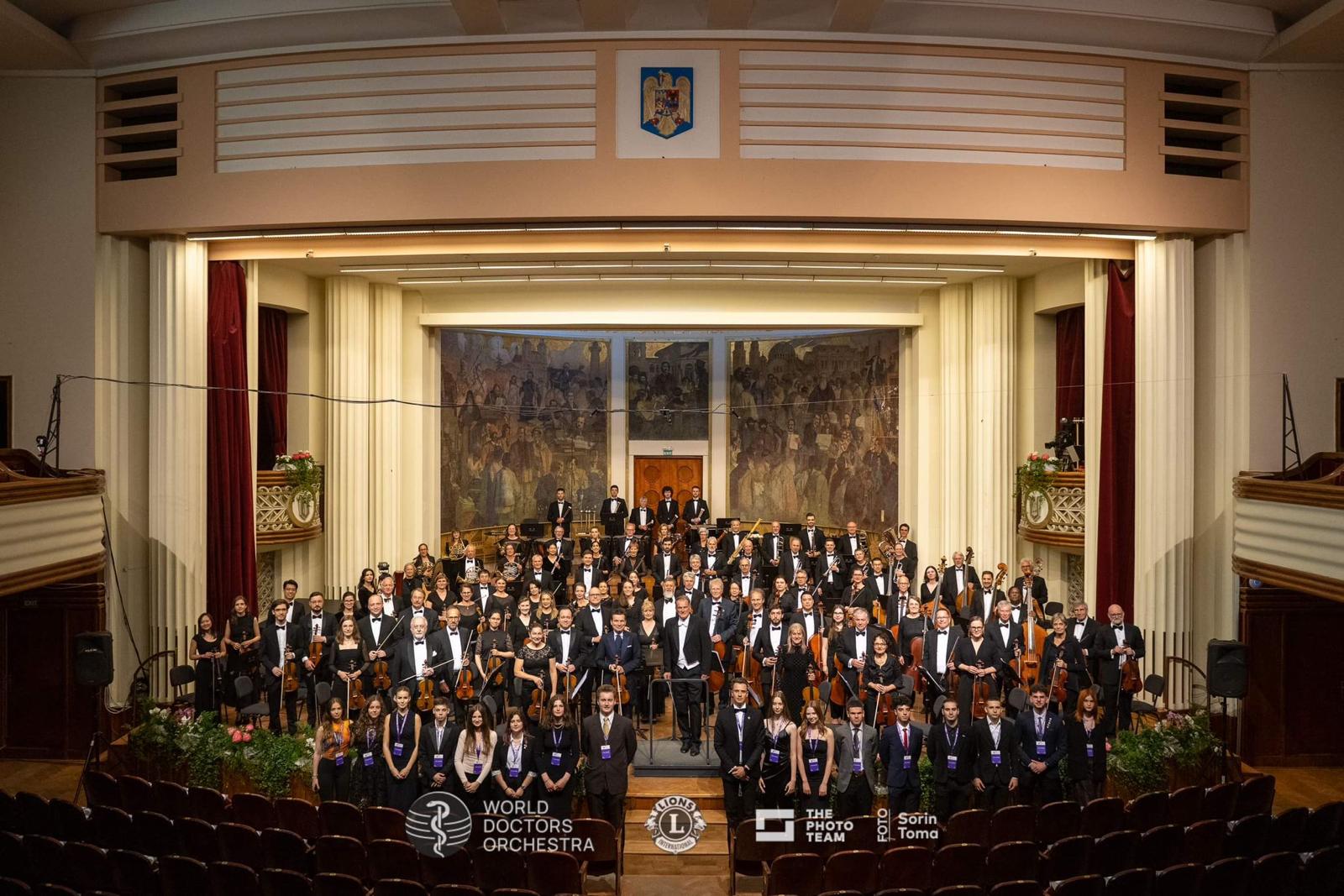 Orchestra group on stage in a grand hall, featuring a decorative mural, with logos of World Doctors Orchestra and partner organizations.