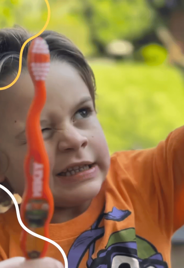 A young child in an orange shirt making a playful face while holding an orange toothbrush outdoors.