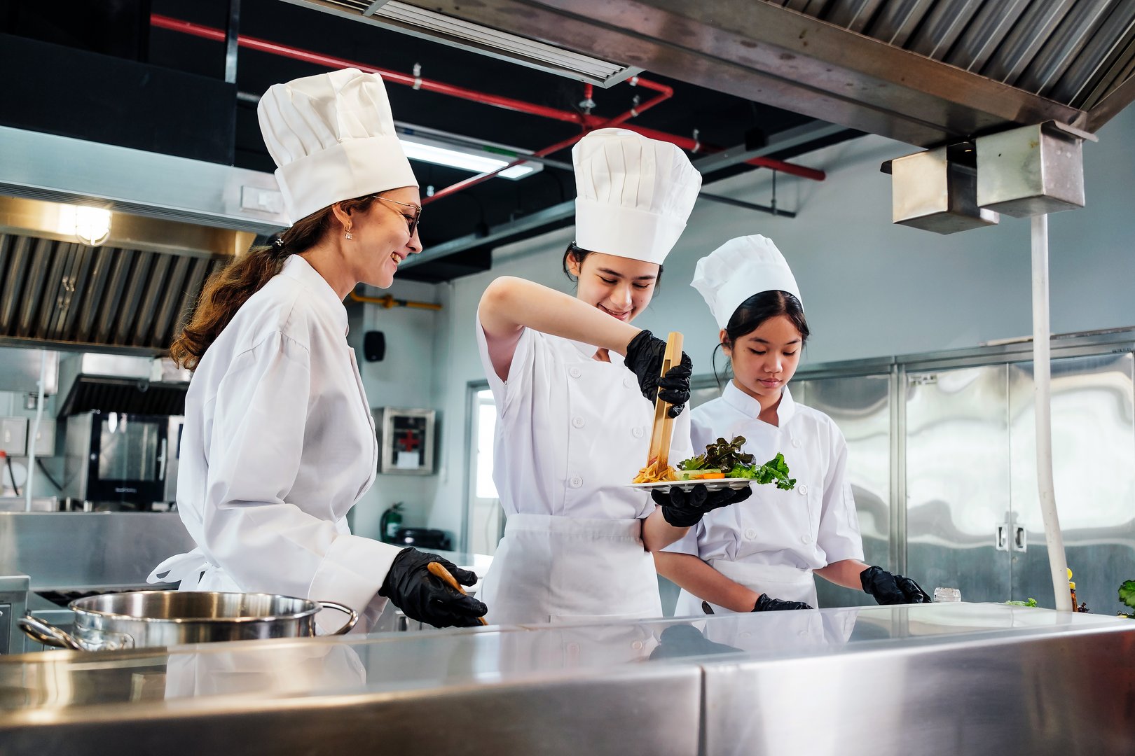 Team of chefs in white uniforms and toques plate gourmet dish in commercial kitchen. Scene shows instructor-led collaboration, dining presentation, stainless workflow, education, teamwork in school.