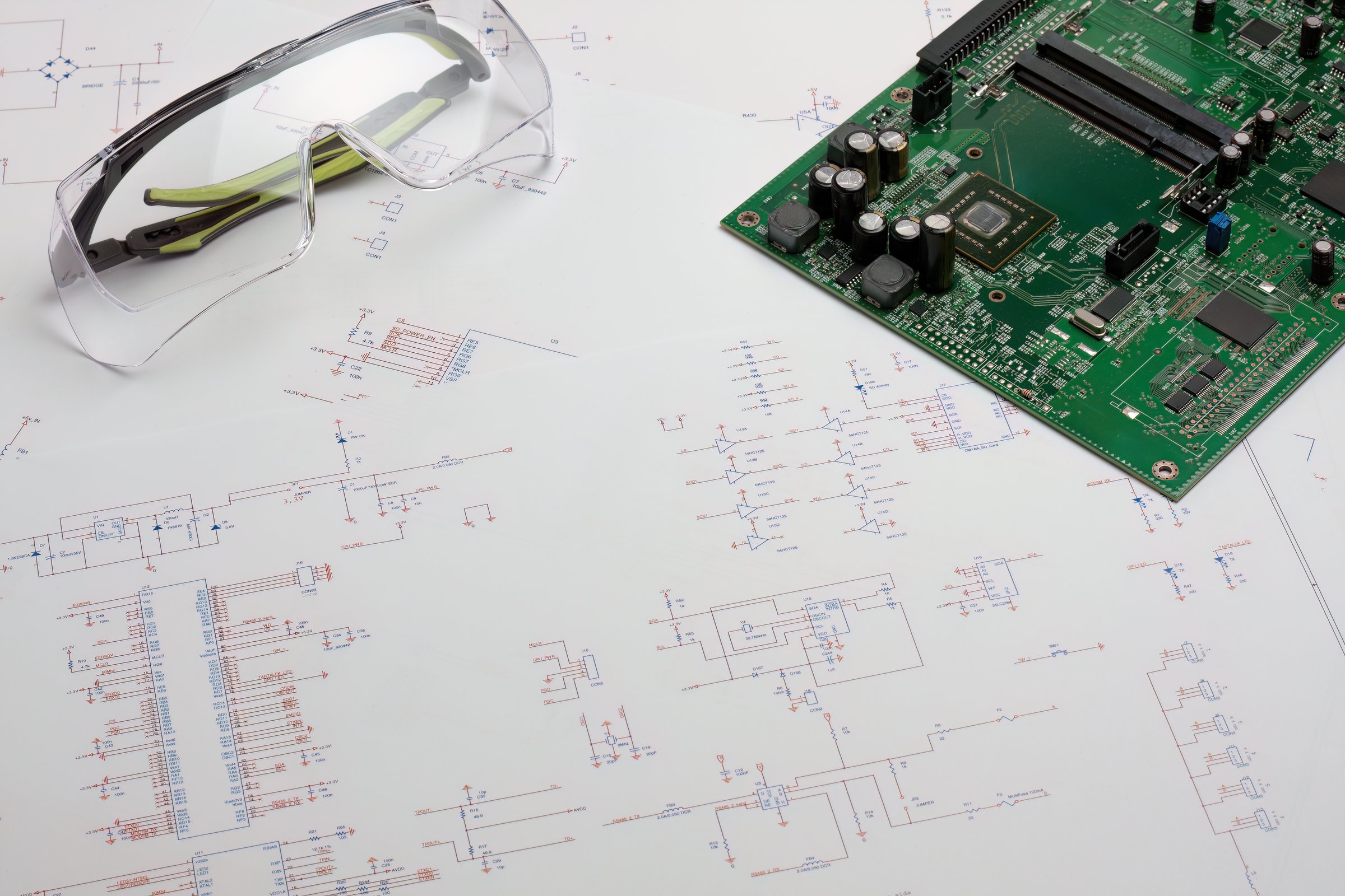 An engineer's desk featuring an electronic blueprints, and a green circuit board and a saftety glasses. The image highlights the process of designing and reviewing electronic components.