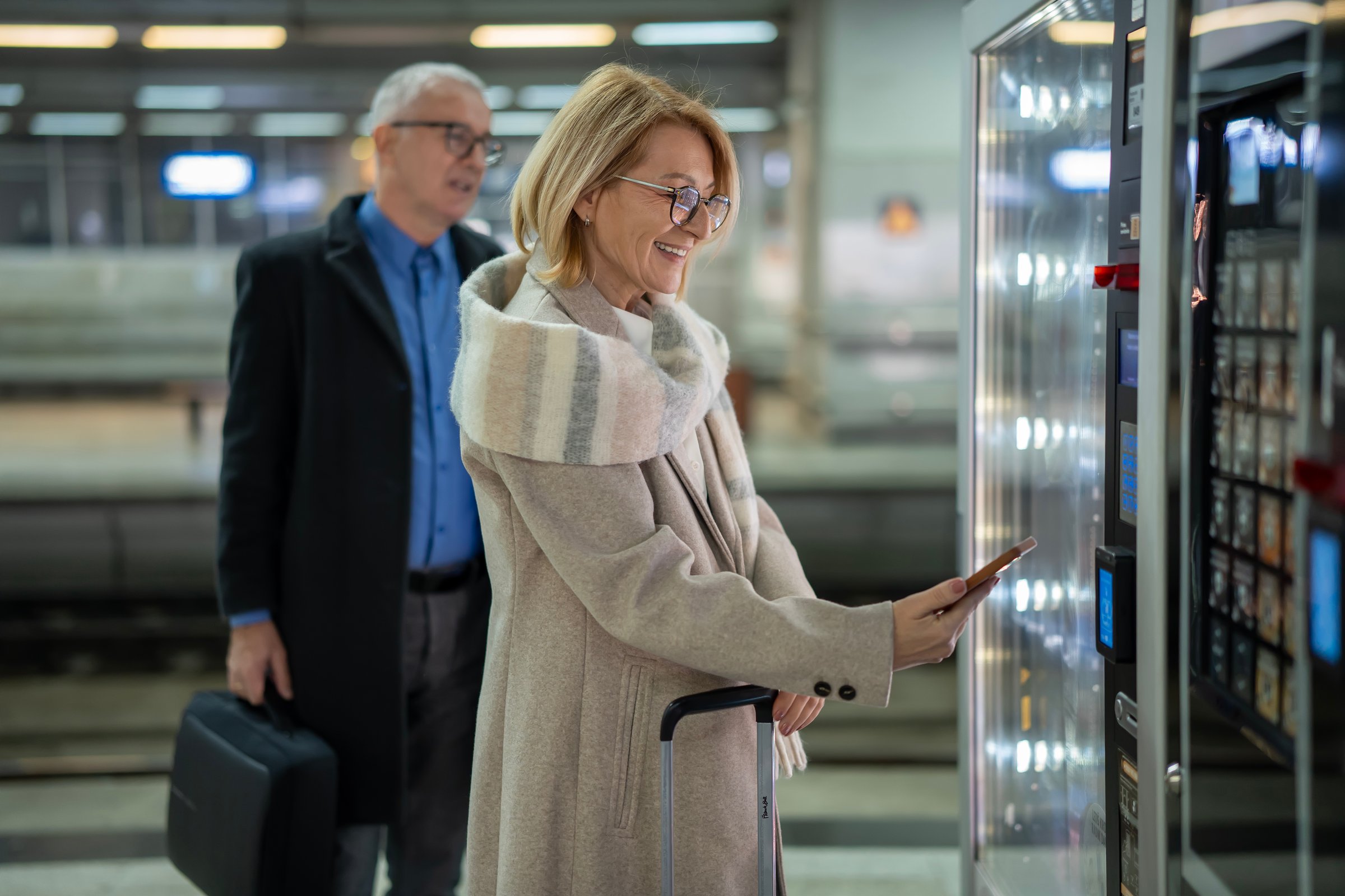 Senior woman smiling as she makes a contactless smartphone payment at a vending machine while traveling with her husband on a train station platform, luggage nearby