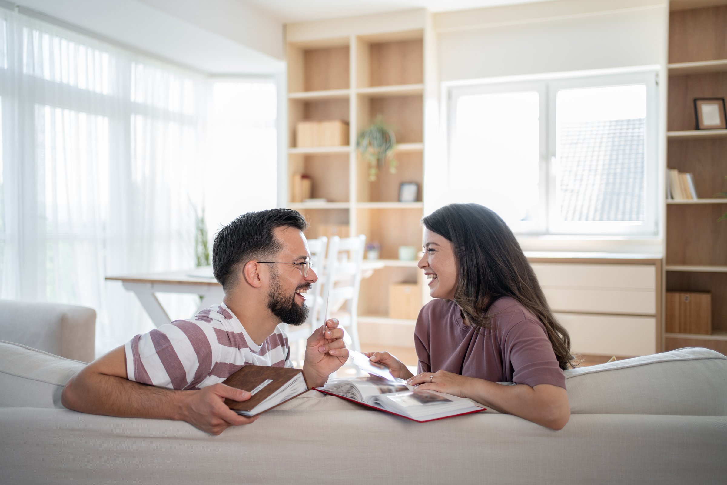 Young couple laughing and sharing joyful moments while browsing through old photo albums, lying comfortably on the sofa in their bright and inviting living room