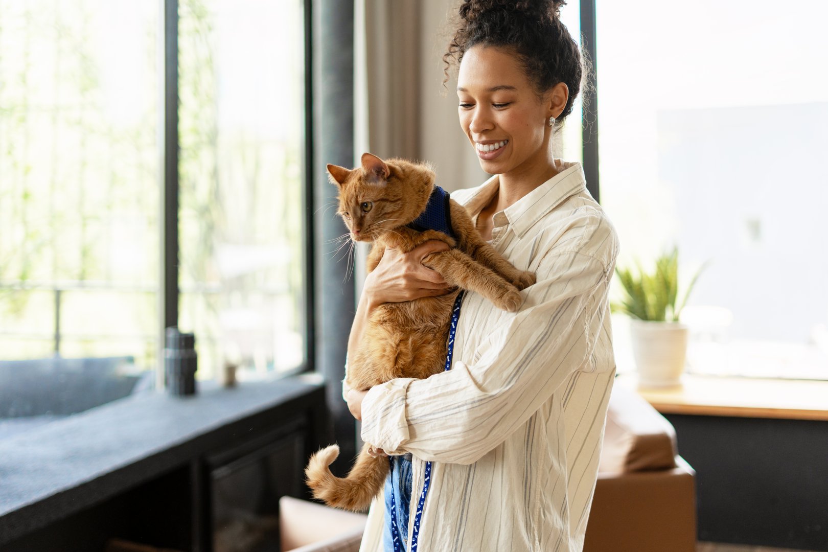 Young adult woman smiling and holding a ginger cat by a window in a bright, modern interior. The scene conveys warmth, happiness, and the bond between pets and owners