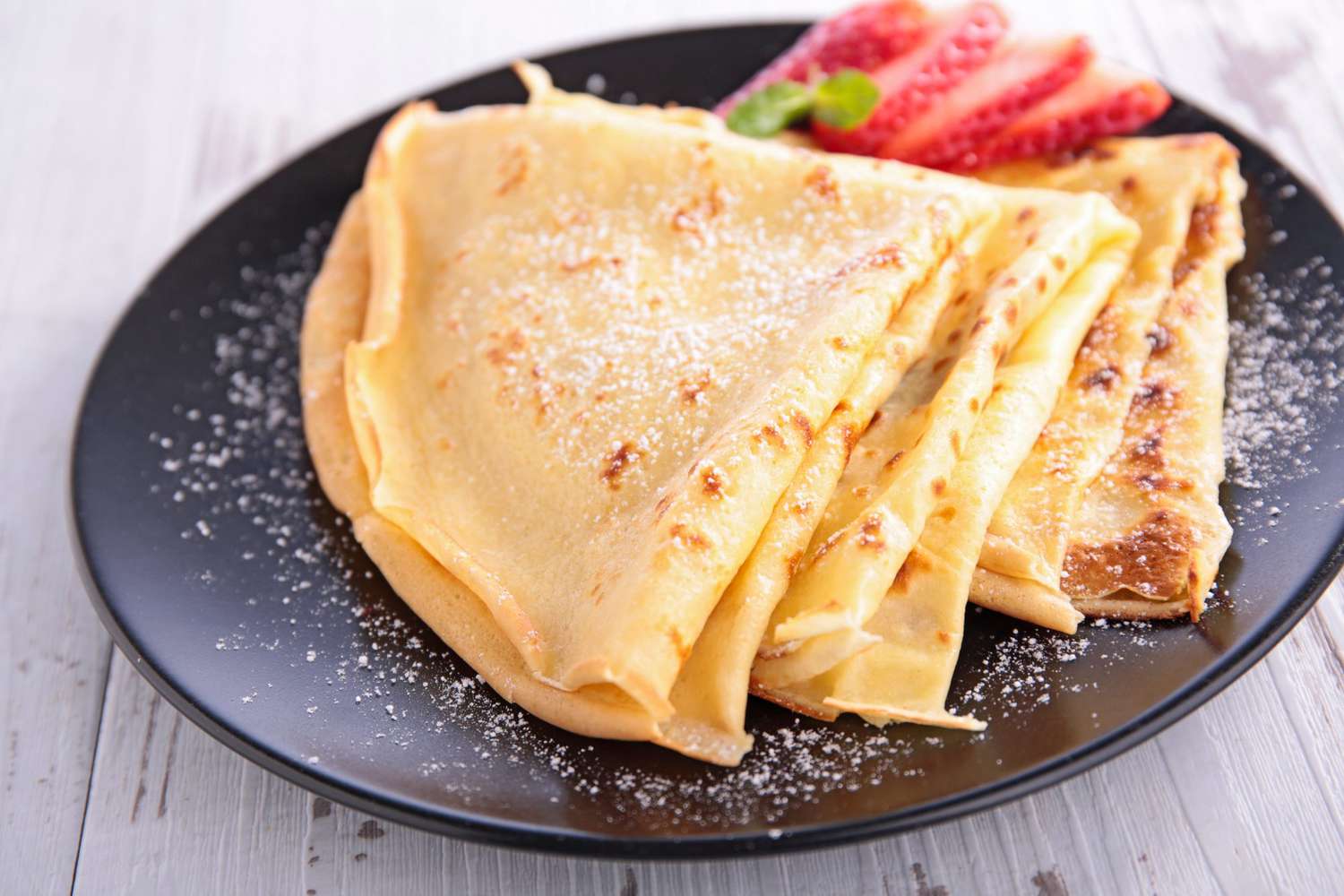 Crepes on a black plate with powdered sugar, garnished with sliced strawberries on a light wooden surface.