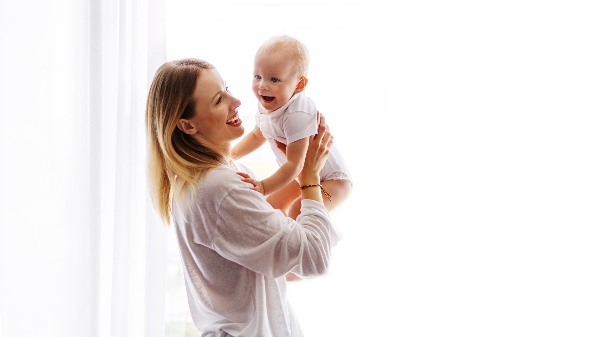 Joyful young mother and baby dressed in white laughing and playing together, tickling and bonding on white background. Beautiful mom and small child in arms