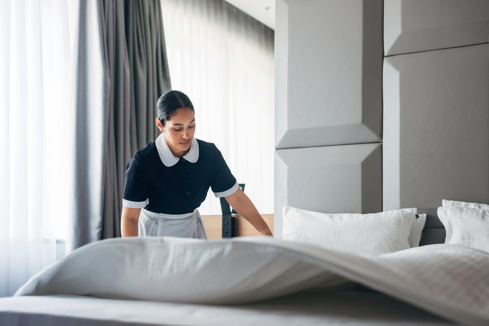 A hotel staff member wearing uniform is tidying up a neatly arranged bed in a stylishly designed room. The bright setting exemplifies cleanliness, hospitality, professionalism, and dedication to providing exceptional guest experiences.