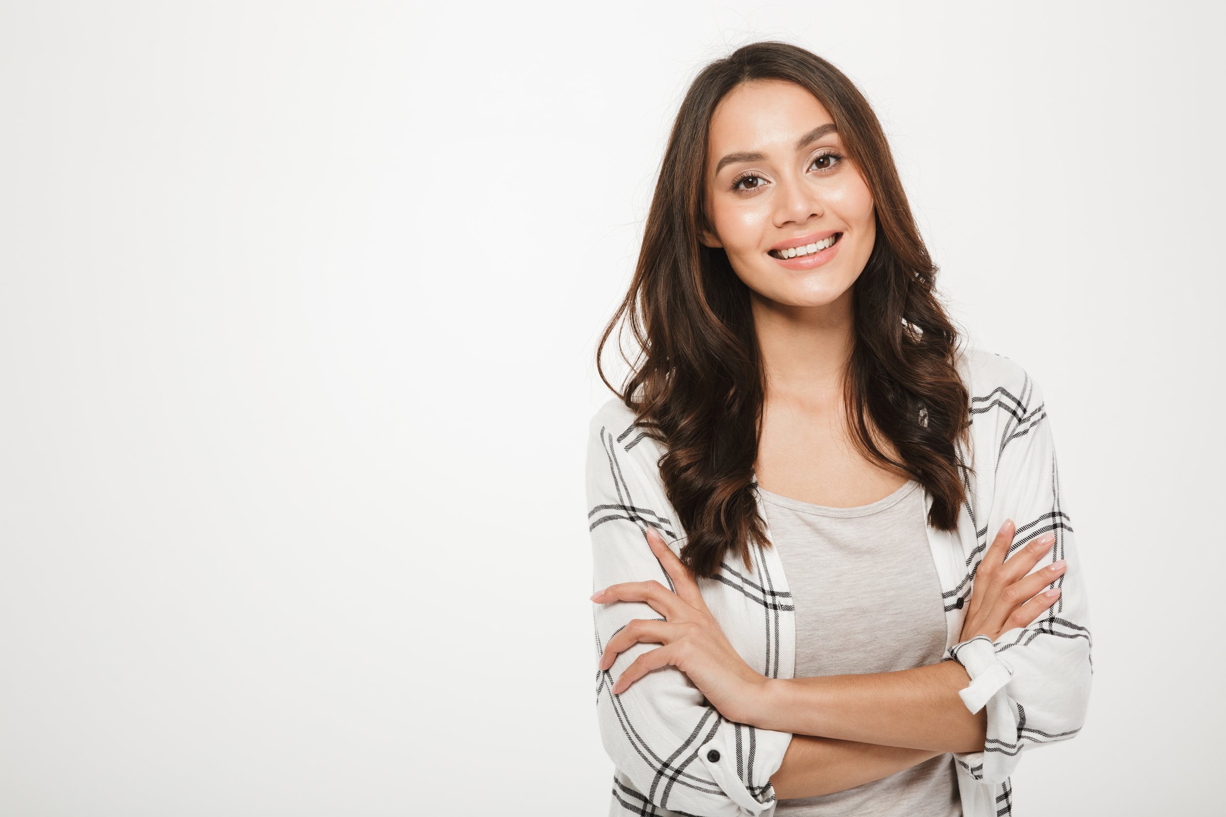 Smiling woman with long brown hair wearing a plaid shirt over a gray top, standing with arms crossed against a white background.