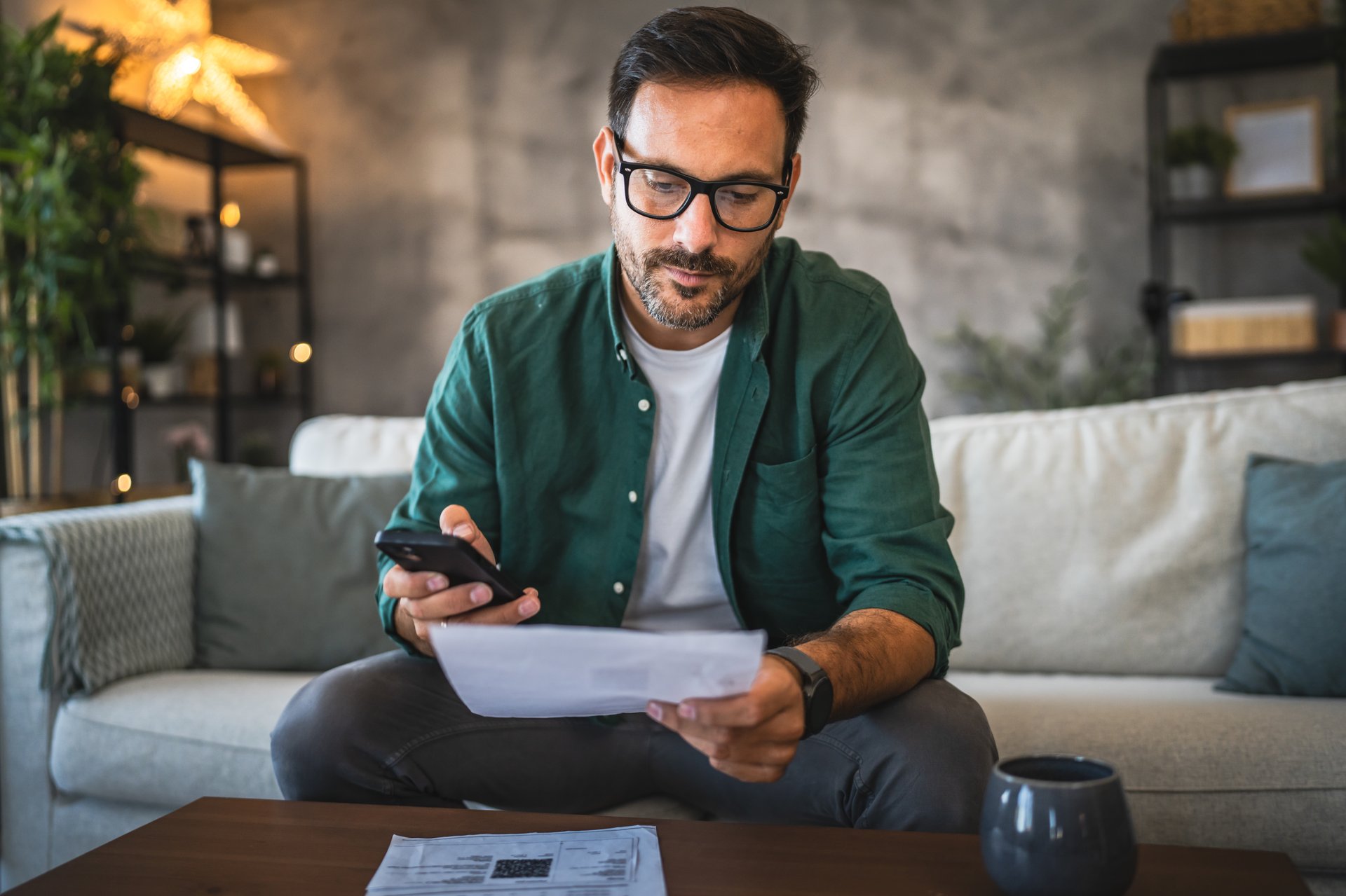 Adult man in casual attire sit on a sofa, holding a bill and a smart phone, with other documents on the table and a coffee mug, focusing on managing his finances