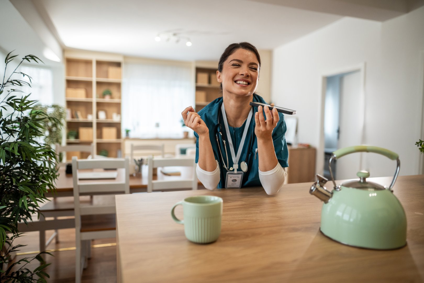 Smiling female nurse wearing scrubs and a stethoscope, standing in a bright home kitchen, enjoying a relaxing coffee break and using her smartphone for a call