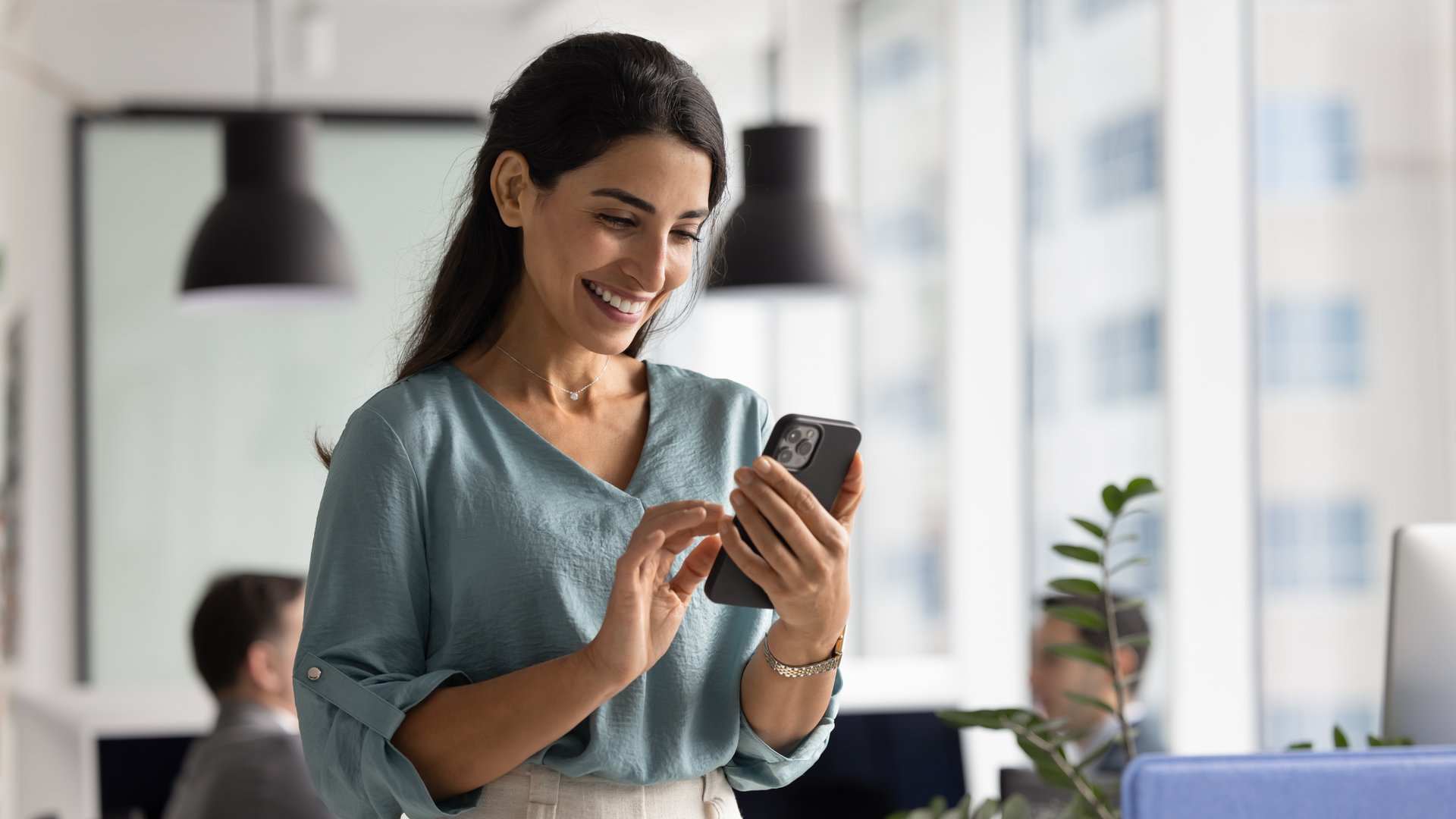 Happy beautiful Hispanic businesswoman using web service on smartphone in office workspace, working on online project on mobile phone, typing on cellphone, smiling. Banner shot