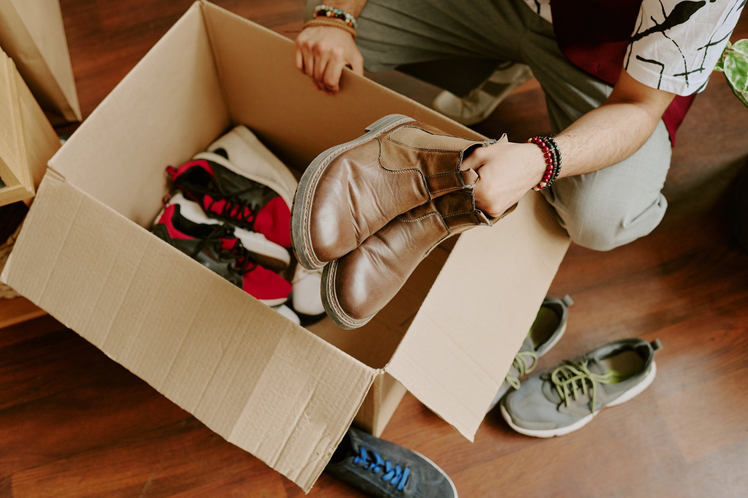Person sorting various pairs of shoes in cardboard box on wooden floor. Different styles of shoes present, with some already inside box and others around it on floor