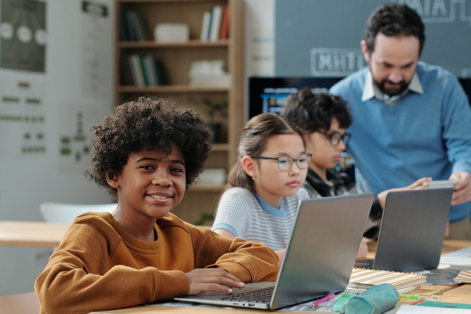 Group of children studying in classroom, focused on tasks on laptops, with teacher overseeing their progress. Classroom bookshelf and posters in background creating educational atmosphere