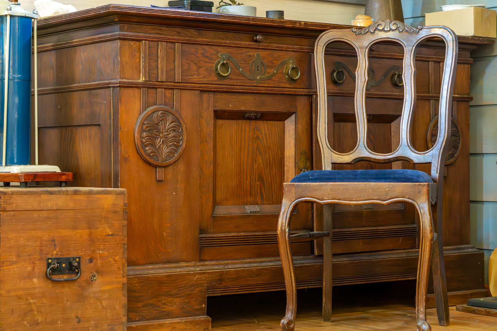 vintage wooden sideboard with carved details and brass handles, accompanied by an elegant wooden chair featuring a blue upholstered seat. Classic furniture likely dating from the late 19th or early 20th century, showcasing traditional craftsmanship.