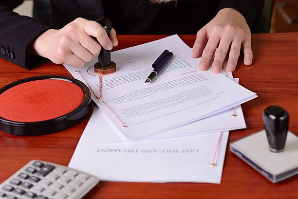 Person stamping a last will and testament document on a wooden desk with a calculator and fountain pen nearby.