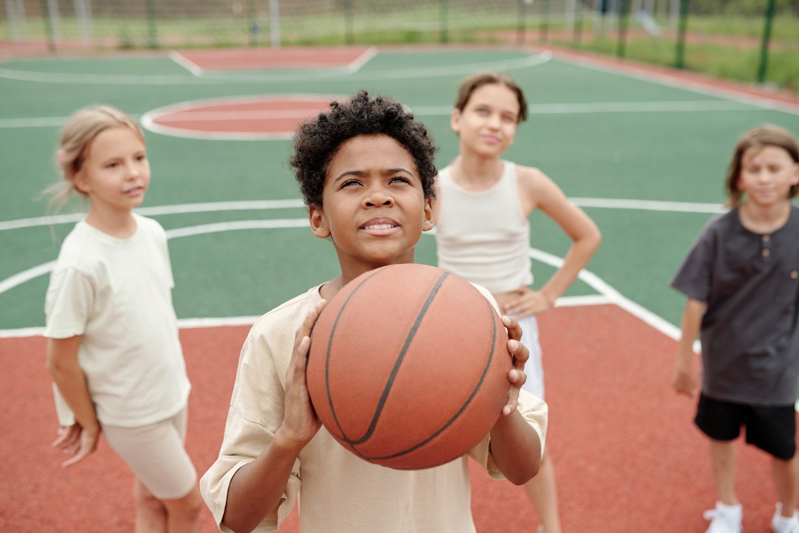 Cute schoolboy with ball looking upwards before throwing it into basket while standing in front of camera against group of classmates