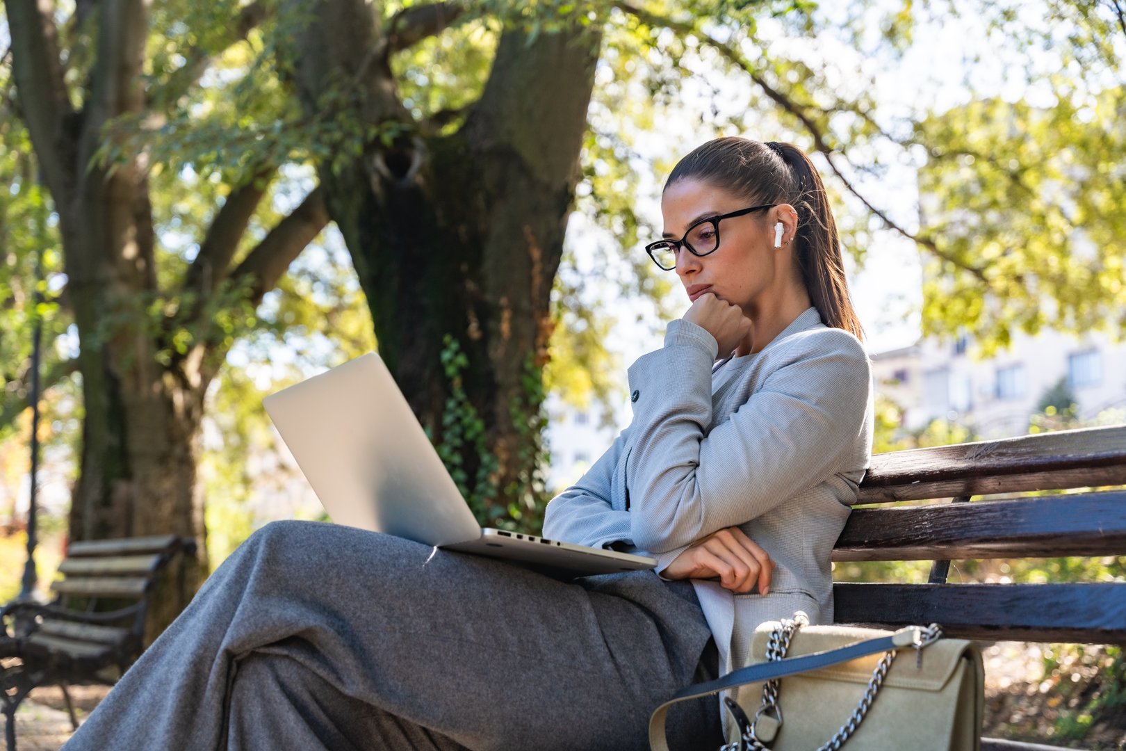 Young successful businesswoman having video call on laptop computer while sitting on the bench in the park. Business female working remote, talking with client on webcam, giving him advice about work.