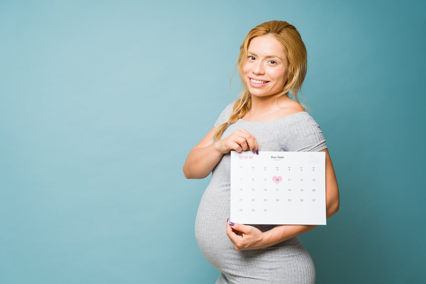 Cute Caucasian pregnant woman in her 30s checking her due date on a calendar while standing against a blue background in a studio