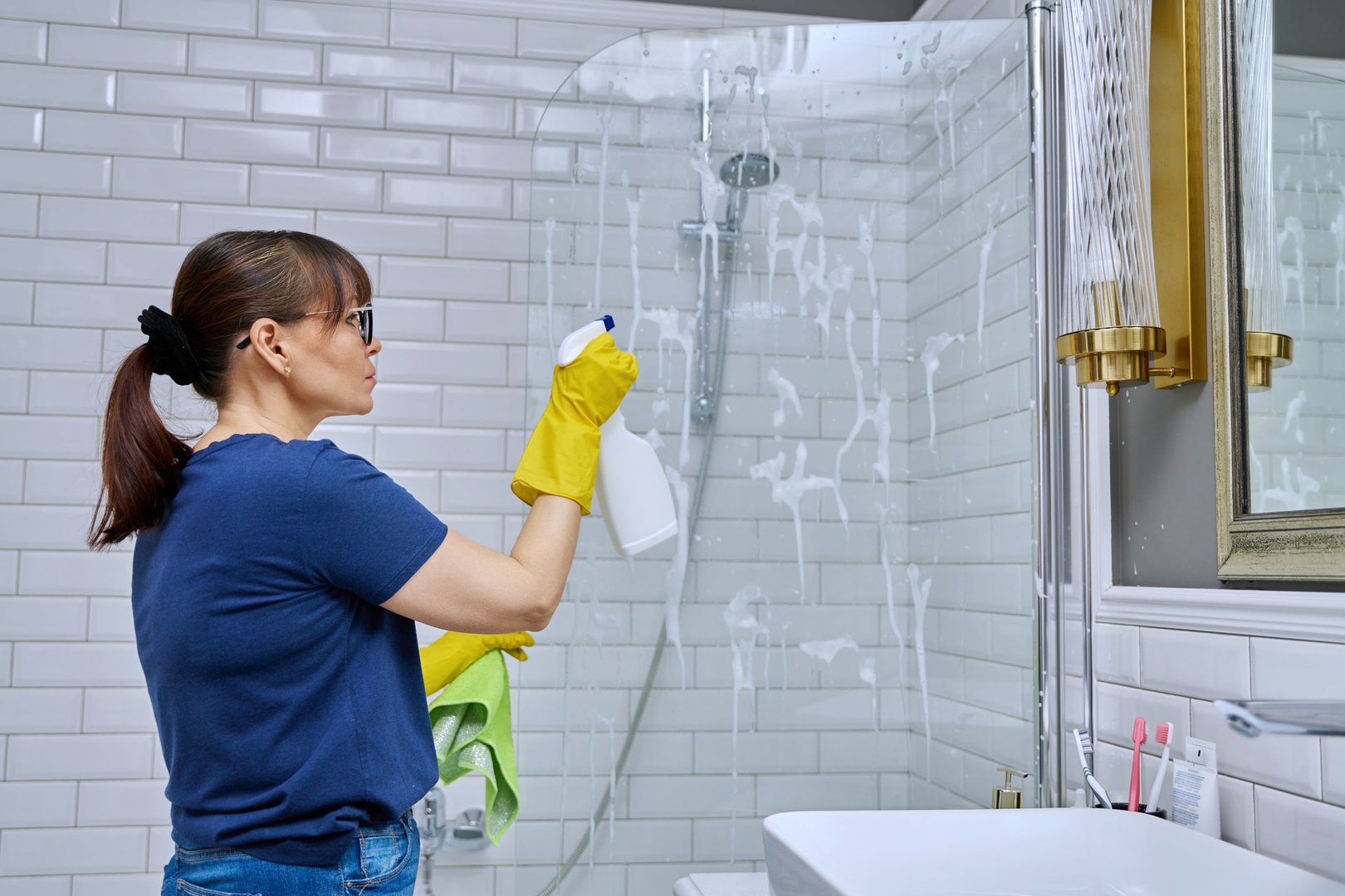Woman cleaning bathroom, washing glass in shower, with detergent spray and professional rag. Housekeeping, housework, housecleaning, cleaning service concept