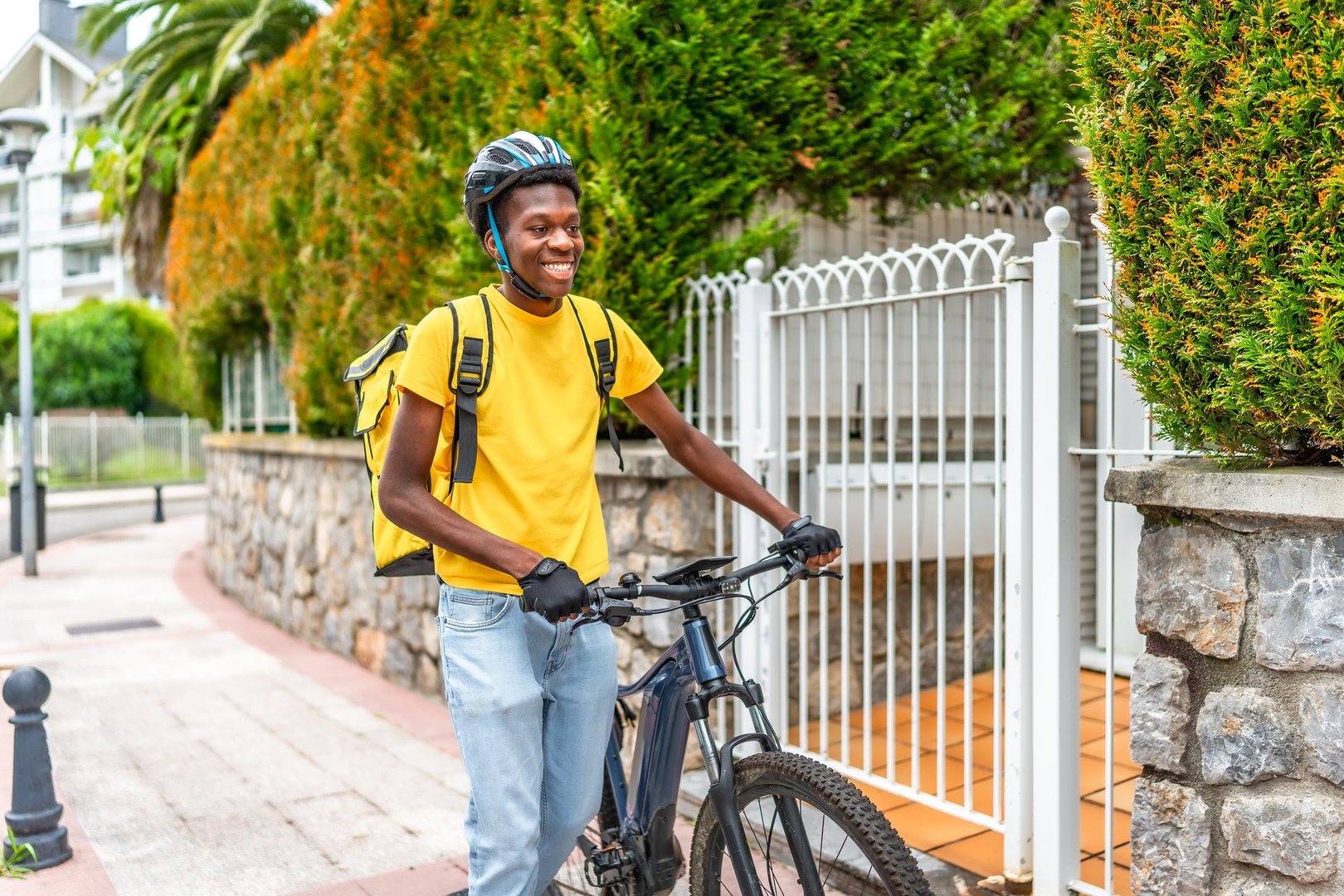 Delivery african man with backpack and helmet holding a bike in a residential area