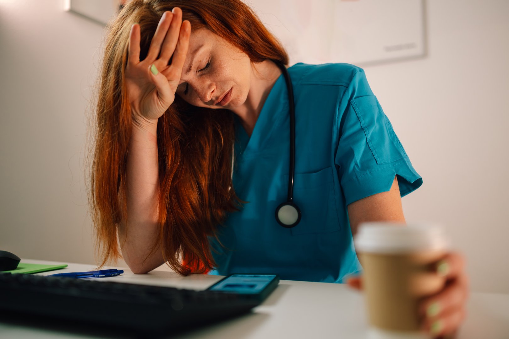 A red-haired nurse in a blue scrub uniform holds a cup of coffee, rests her head on her hand, and studies her phone at a desk, displaying signs of stress and busyness in her professional environment.