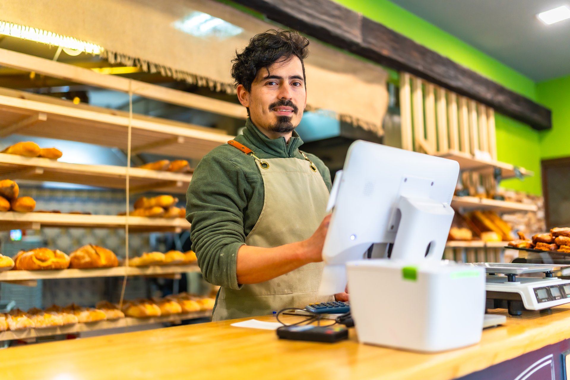 Latin friendly salesman in apron using computer stranding in the counter of a bakery shop