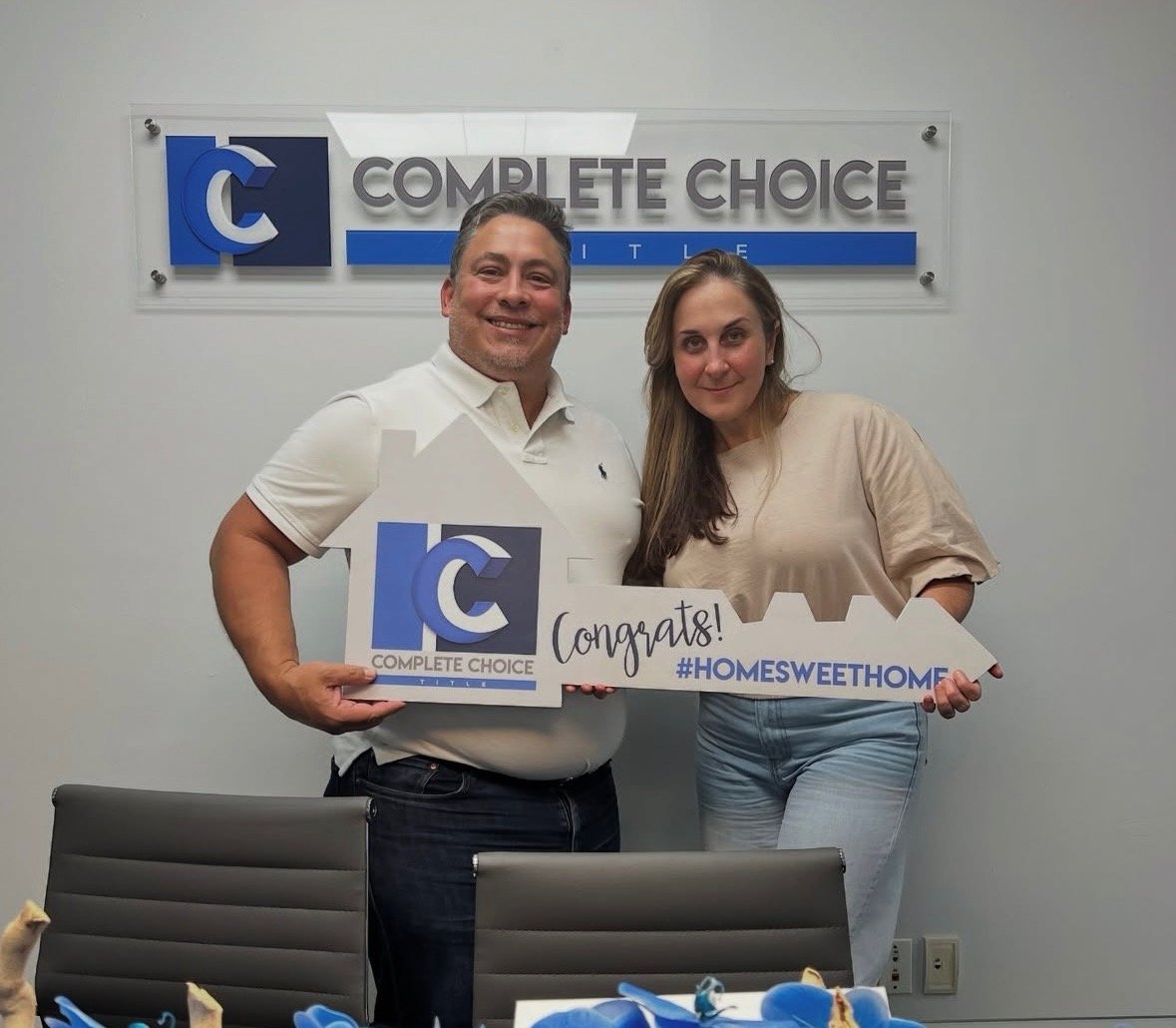Two people smiling, holding a house-shaped sign in an office. Sign reads Congrats! #HOMESWEETHOME with Complete Choice Title logo.