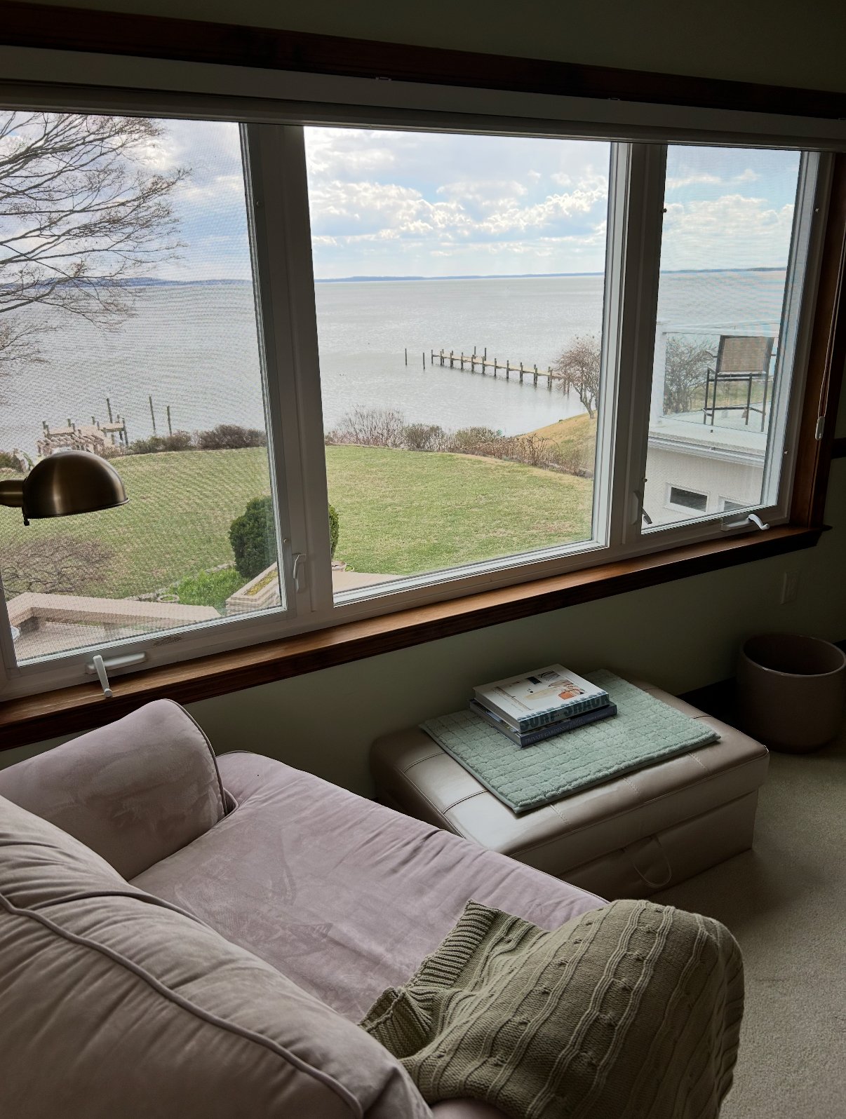Room with a beige couch and ottoman, large window view of a lawn, pier, and waterfront.