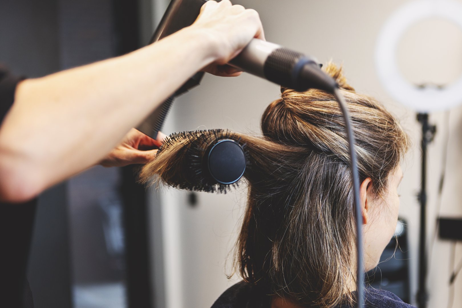 Hairdresser blow drying hair of woman with round brush in modern beauty salon, styling sectioned strands. Close up professional hairstyling