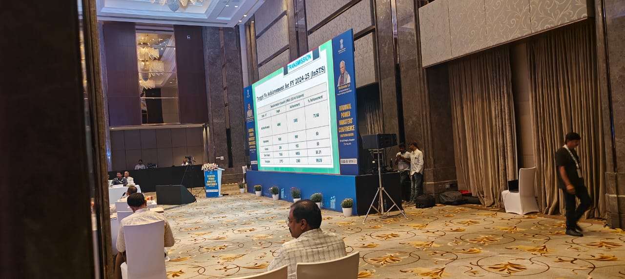 Conference room with people seated, large screen displaying data, and a speaker standing near the stage.