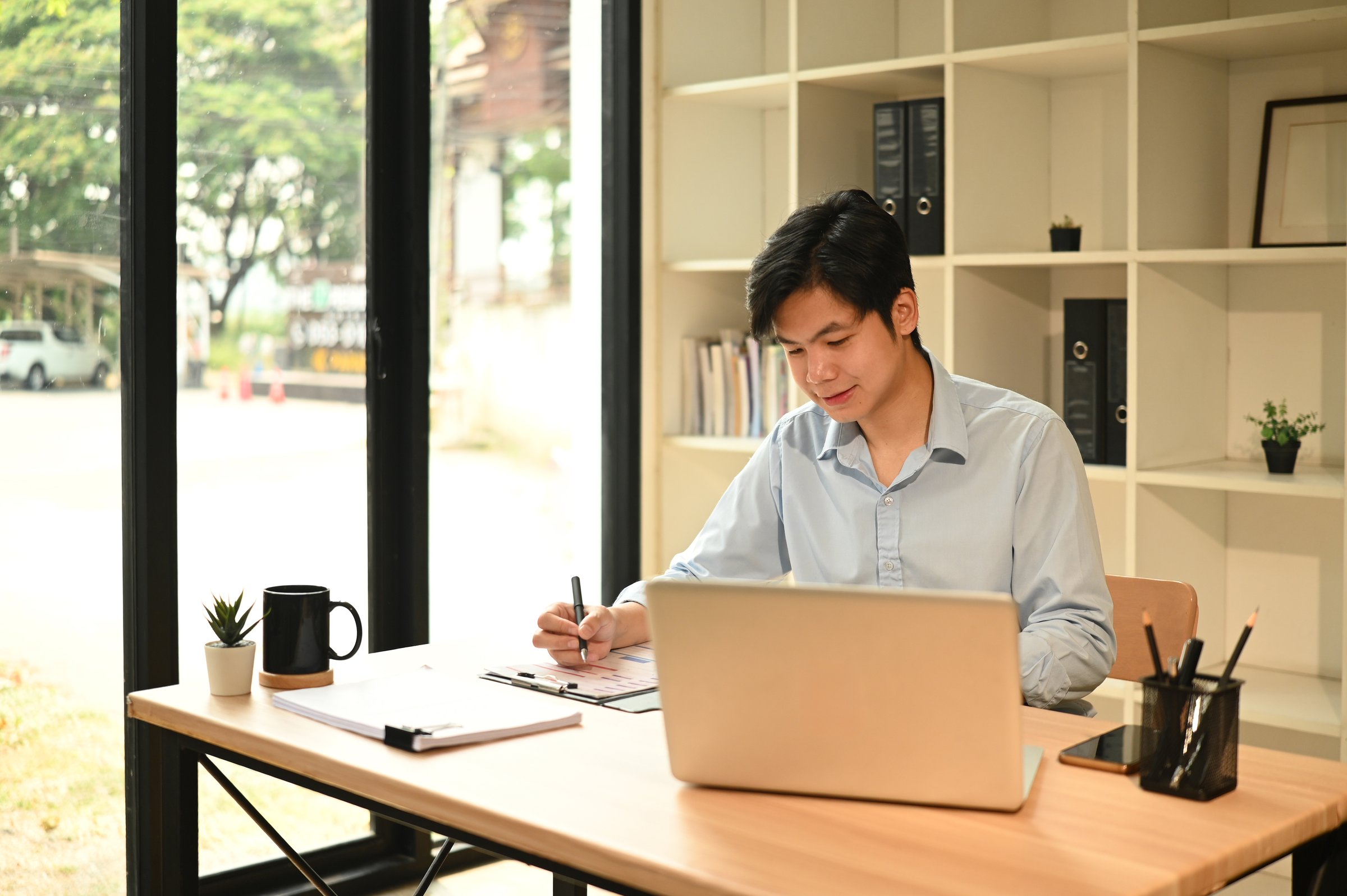 Confident Professional Asian man working on a laptop at an Office Desk in Modern Workspace.