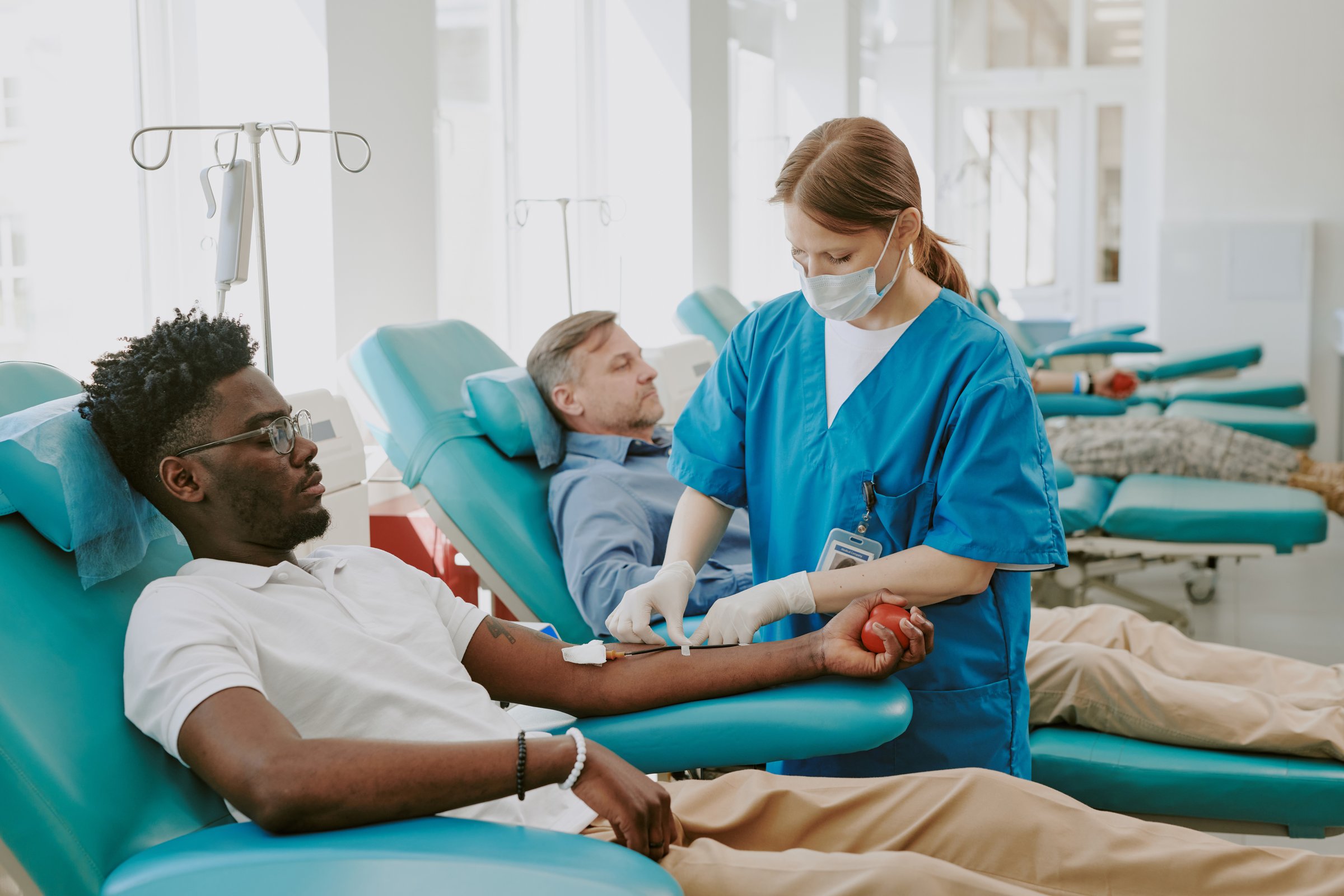 Young Black man donating blood while female healthcare worker preparing needle, middle aged Caucasian man reclining in background donating blood, medical chairs and equipment visible