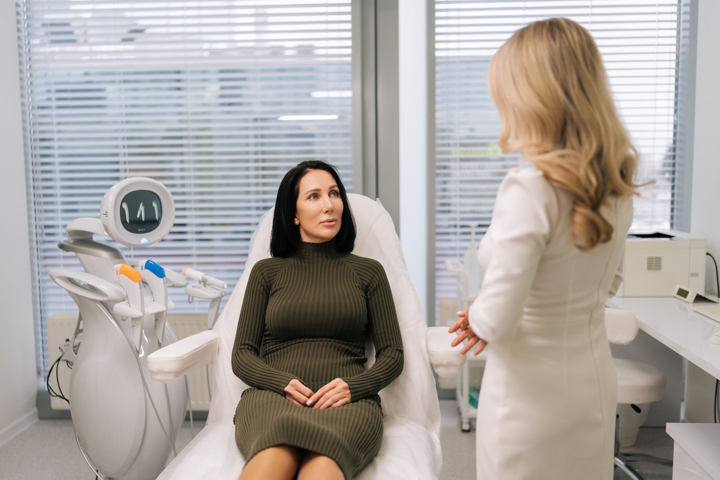 Back view of cosmetologist in white uniform consulting adult middle-aged client about ultrasound-based skin care and non-surgical body contouring in modern clinic setting for antiaging treatments.