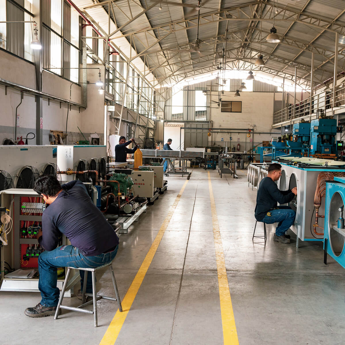 Workers assembling equipment in a spacious factory with high ceilings and industrial machinery.