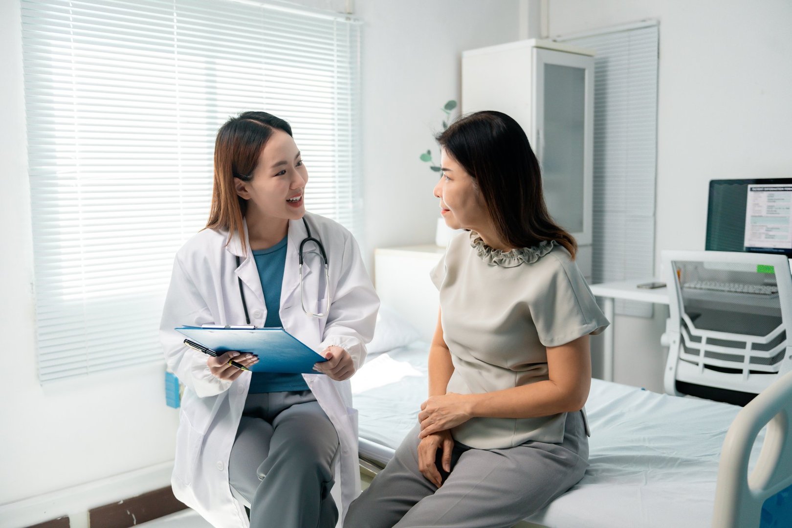 Young female doctor holding a clipboard and talking to a mature female patient sitting on a hospital bed, discussing medical records and treatment plans in a bright and modern hospital room