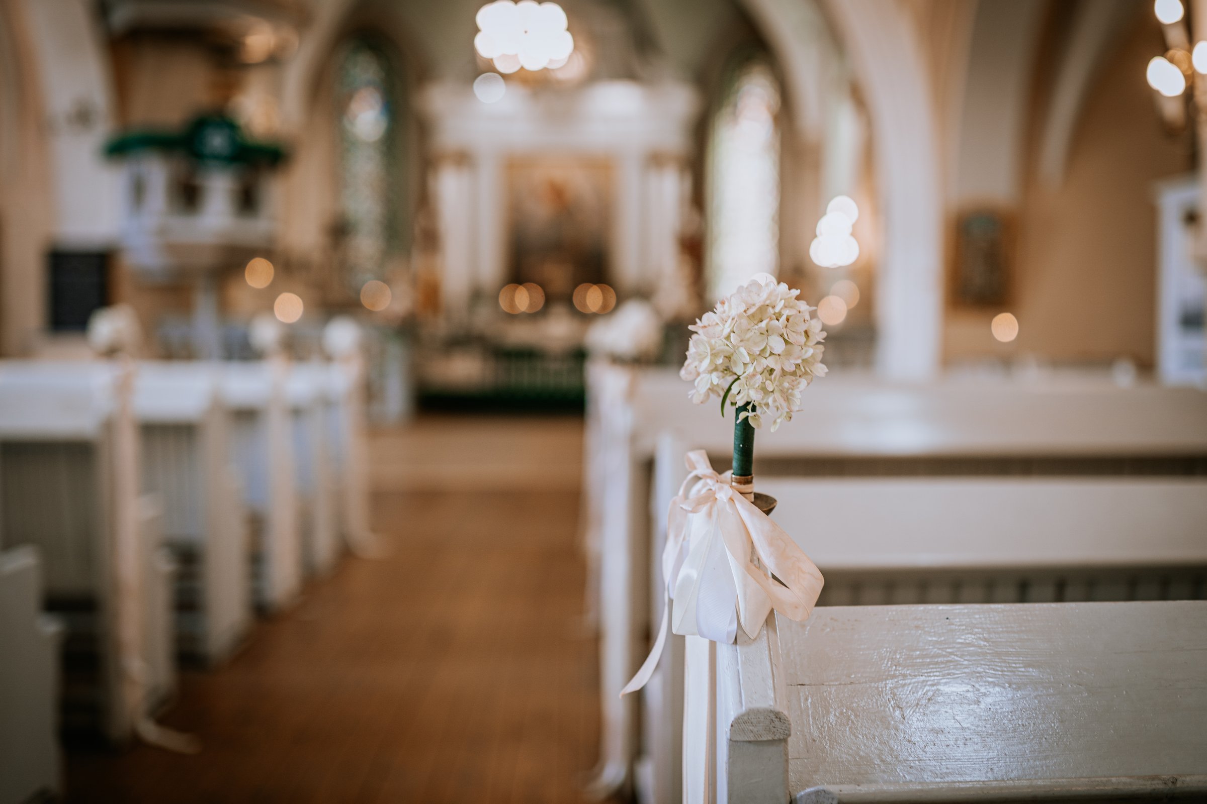 Valmiera, Latvia - August 9, 2024 - Close-up of a white floral wedding decoration with ribbon on a church pew, with a blurred altar and soft lighting in the background.