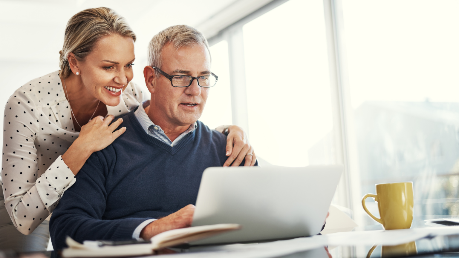 Smiling woman and man at a desk using a laptop, with a notebook and coffee mug. Bright, modern office setting.