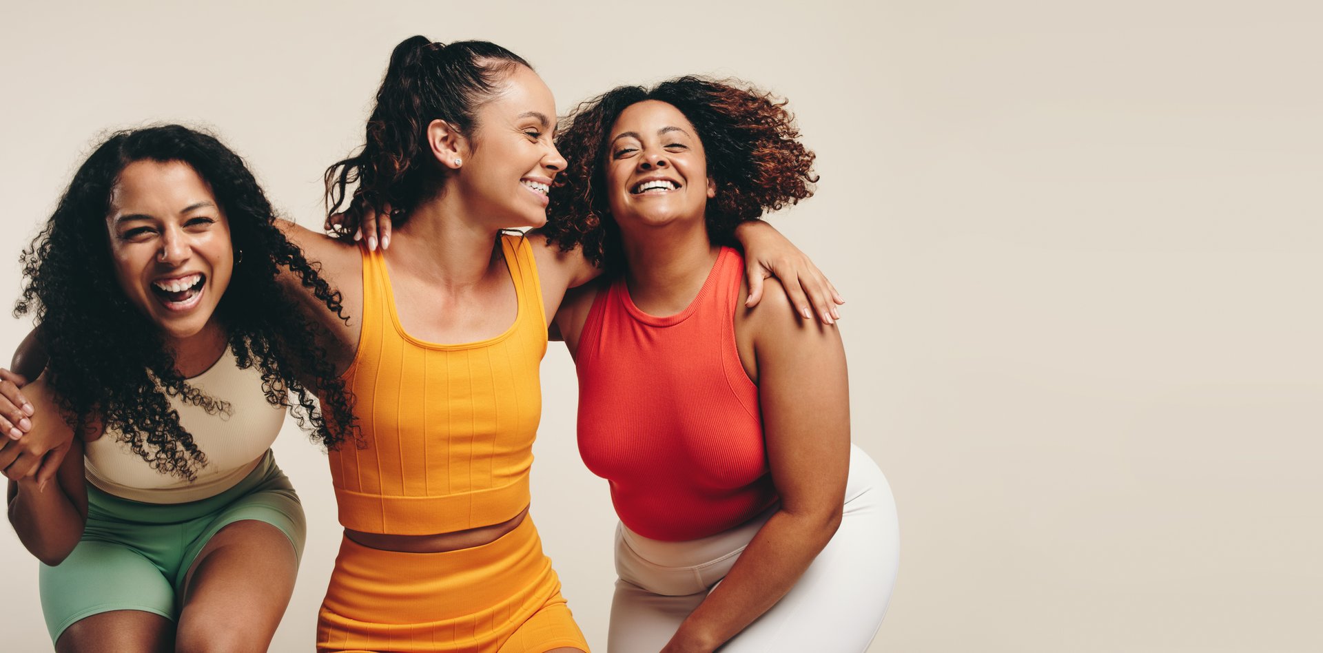 Group of three young, diverse female athletes celebrate their healthy and active lifestyle in a sports studio, smiling and laughing together while wearing sporty fitness clothes.