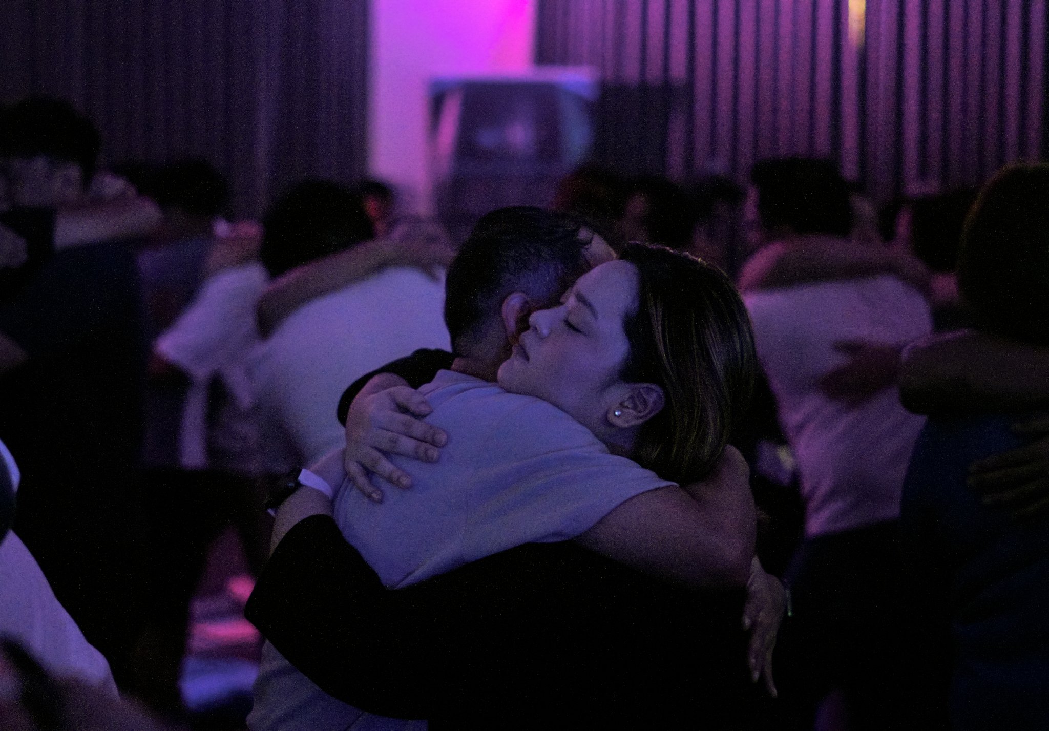 People embrace in a dimly lit room with purple lighting, creating an intimate atmosphere.