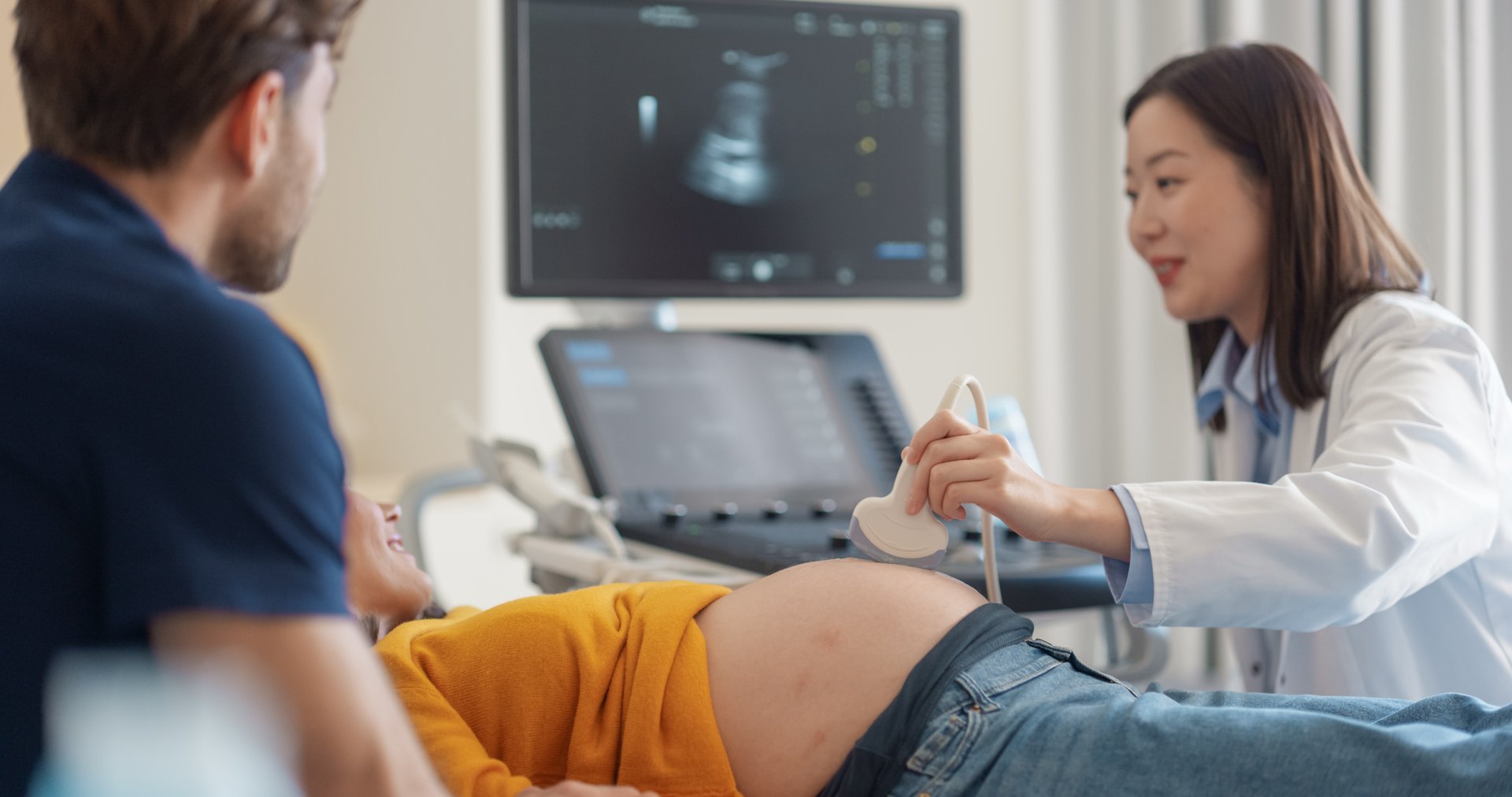 Asian Obstetrician Conducts Prenatal Ultrasound for Smiling Expectant Mother in Yellow Top. Husband Observes as Doctor Explains Fetal Development Using Advanced Imaging Technology in Modern Hospital