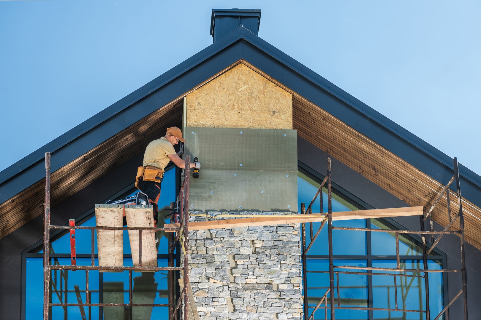 A construction worker, wearing safety gear, stands on scaffolding, using a drill to install stone veneer on a modern home. The home features a black roof and large windows. The construction is in progress, with visible framing and scaffolding.