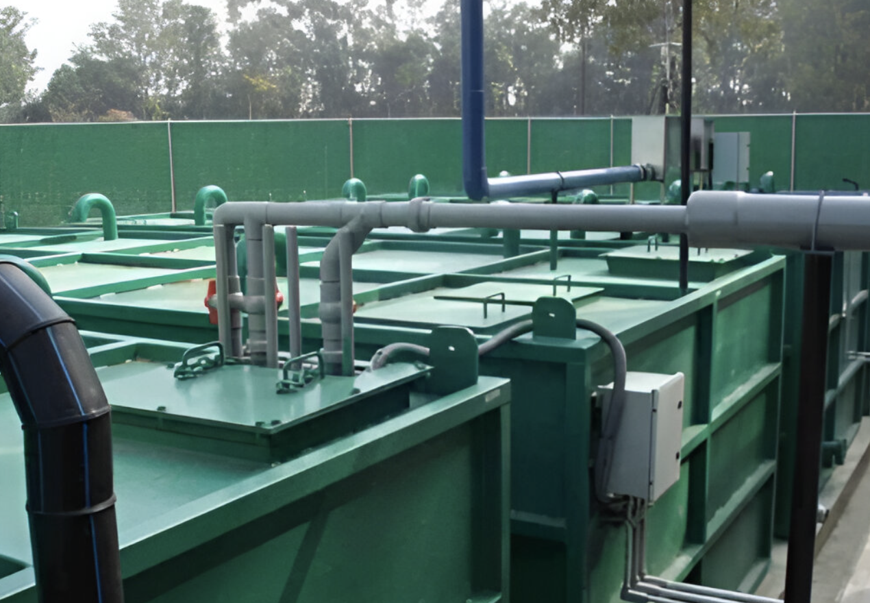 Green industrial tanks connected by pipes in an outdoor area, surrounded by trees and a green fence.